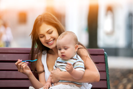 Baby feeding spoon by mother in park outdoor. Weaning in first weeks. Mom and eating child summer on bench. Good appetite by baby food with foster mother . Color tone on shiny sunlight background.