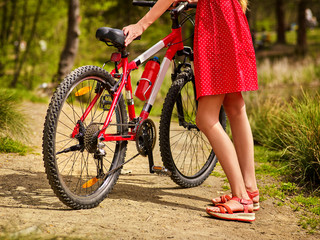 Fototapeta na wymiar Bicycle and low section female legs by girl wearing red polka dots dress rides into park. Girl in ecotourism. Cycling is good for health. Cyclist is unrecognizable.