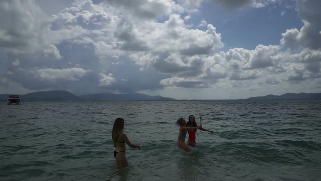 Three beautiful young happy women in bikini's enjoying their holiday on the sandy beach on tropical paradise island. One girl with selfie-stick doing photos in the water - video in slow motion