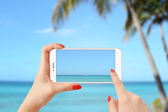 Girl on the beach taking picture of sailing boat on the sea with smartphone
