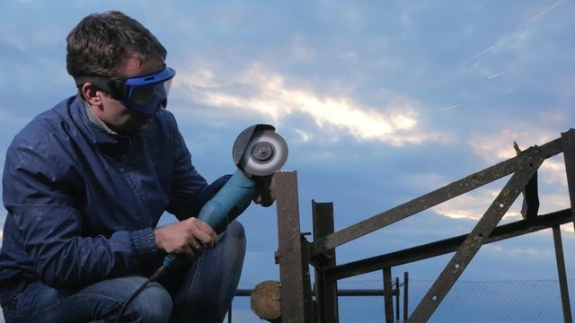 A young man polishes a metal angle grinder at sunset. Beautiful sky behind the worker.