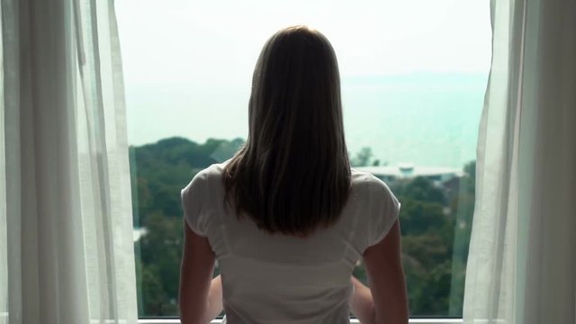 Woman in white t-shirt unveiling curtains and looking out of window. Enjoying the sea view 100 fps