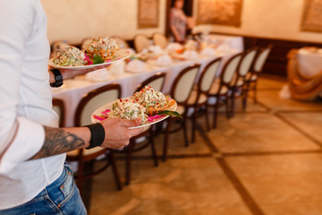 Waiter with the tattoo carrying plates with meat dish to the tables.
