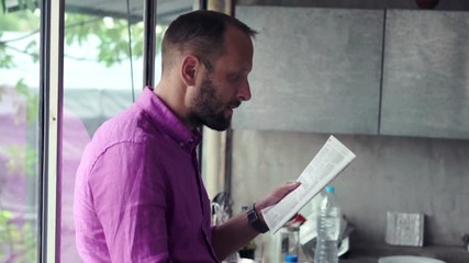 Young man reading newspaper and drinking tea sitting by the window in the kitchen
