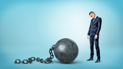 A small sad businessman looking down to a giant iron ball and chain on blue background.