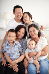 Portrait of big Asian family posing with little kids at home sitting on sofa, all smiling happily and looking at camera