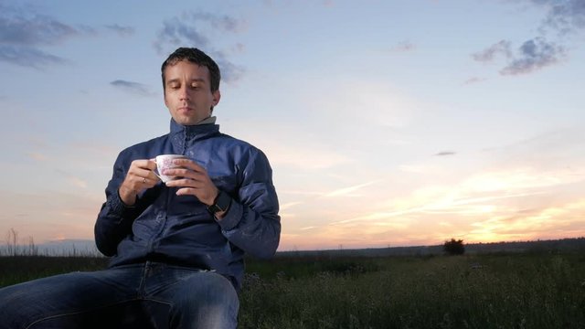 A young man drinks a hot drink from a mug at sunset. Beautiful sky behind.