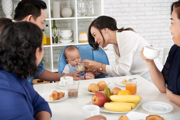 Portrait of happy Asian family at breakfast, mother feeding baby boy at dining table and smiling