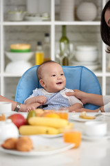 Portrait of adorable Asian baby boy smiling, sitting in little chair with family at dining table during breakfast
