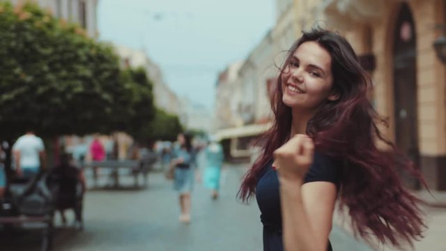 Lovely young brunette woman in a black casual wear walks down the central city street, turns and smiles right to the camera, posing and playing with her long hair. Being emotional, cheerful mood.