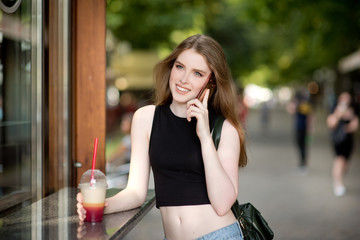 Fototapeta premium Young beautiful stylishly dressed girl is walking along the streets of the city, drinking juice, coffee, talking on mobile phone