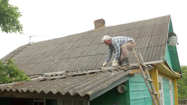 An elderly man is fixing the roof himself. Old wooden house in slate
