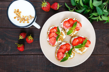 Mini sandwiches with cottage cheese, fresh strawberries, decorated with mint leaves on rye bread and a mug of yogurt on a dark wooden background. Top view. Proper nutrition. Healthy food.Dietary menu