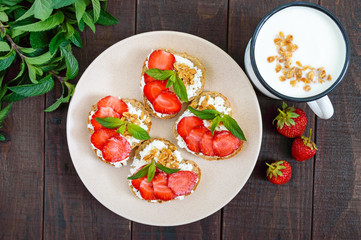Mini sandwiches with cottage cheese, fresh strawberries, decorated with mint leaves on rye bread and a mug of yogurt on a dark wooden background. Top view. Proper nutrition. Healthy food. Dietary menu