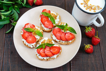 Mini sandwiches with cottage cheese, fresh strawberries, decorated with mint leaves on rye bread and a mug of yogurt on a dark wooden background. Top view. Proper nutrition. Healthy food. Dietary menu