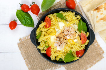 Fusilli pasta, fried eggs and tomatoes. Serve on a cast-iron frying pan on a white wooden background. Top view.