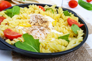 Fusilli pasta, fried eggs and tomatoes. Serve on a cast-iron frying pan on a white wooden background. Close up