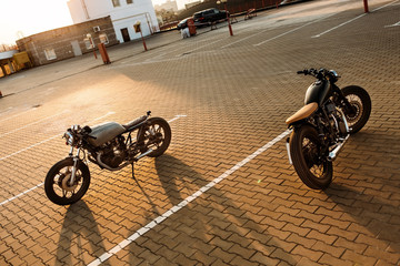Two vintage custom motorcycle caferacer silver and black motorbikes directed in opposite directions on empty rooftop parking lot during sunset. Hipster lifestyle. Confrontation of urban styles.