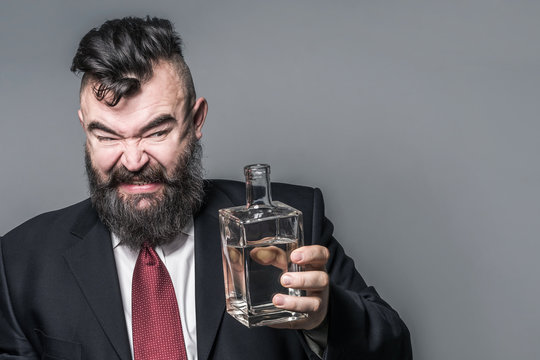 Adult bearded man in suit looking at a bottle of alcohol with disgust on a gray background