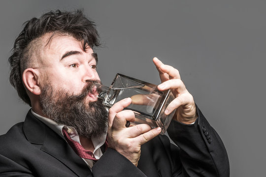Adult bearded man in suit drinks alcohol from a glass bottle on a gray background