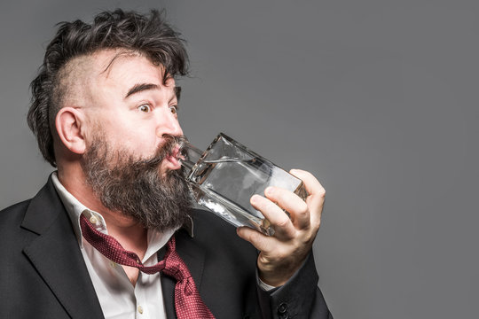 Disheveled bearded man in suit drinks alcohol from a glass bottle on a gray background