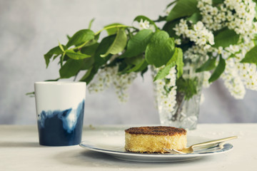 Piece of homemade semolina cake on plate, cup of tea and vase with blooming bird-cherry tree branches on grey concrete background. Spring summer breakfast. Selective focus. Toned image 