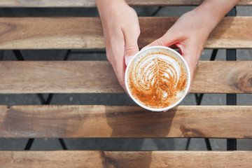 Woman holding a paper cup with coffee. Coffee to go. Tasty hot beverage on wooden table in sunny day. Outdoors meal. Flat lay, top view.