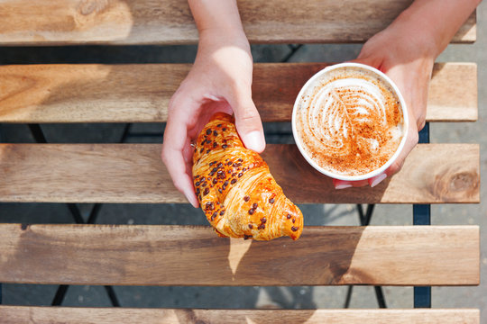 Woman Holding A Paper Cup With Coffee And Chocolate Croissant. Coffee To Go. Tasty Hot Beverage On Wooden Table In Sunny Day. Outdoors Meal. Flat Lay, Top View.