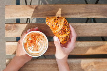 Woman holding a paper cup with coffee and chocolate croissant. Coffee to go. Tasty hot beverage on wooden table in sunny day. Outdoors meal. Flat lay, top view.
