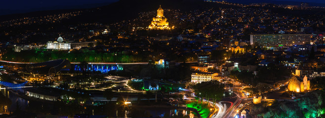 Fototapeta na wymiar Night panorama view of Tbilisi, capital of Georgia country. Metekhi church, Holy Trinity Cathedral (Sameba) and Presidential Administration at night with illumination and moving cars.