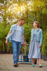 Naklejka premium Man and woman professional gardeners pulling the wagon with potted tree in the garden together and smiling.