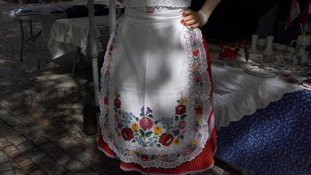 Young slim woman wearing national traditional Hungarian clothes and footwear is standing by souvenir stall 