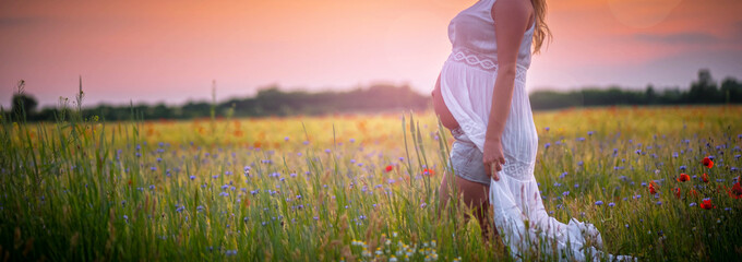 Im 9. Monat schwangere Frau geht im Sonnenuntergang in einer Blumenwiese