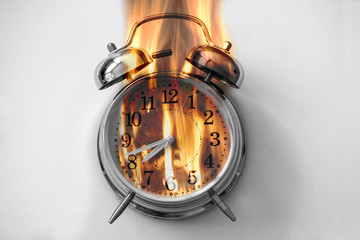 An old alarm clock is on a white desk and big fire flames are coming from it: time is burning
