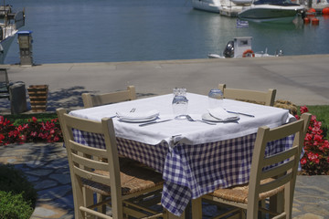 Greek empty outdoor taverna restaurant chairs and table by the sea.
A Greek traditional tavern with white tablecloth, wooden chairs, dishes, forks knifes and glasses on a sunny day.
