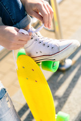 Close up of sexy woman with penny board skateboard in jeans and white sneakers sit on railings and tying laces. Modern urban hipster girl have fun. Good sunny summer day for lifestyle skateboarding.