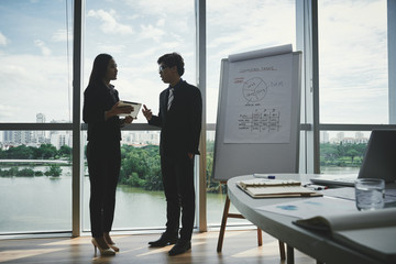 Profile view of confident Asian colleagues discussing joint project while standing at panoramic window in modern meeting room, picturesque cityscape on background