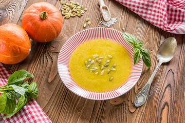 cream soup from pumpkin and other vegetables decorated by basil leaf and pumpkin seeds, basil, pumpkins fruits, salt, pumpkins  seeds  on wooden table with silver spoon on red squared  napkin