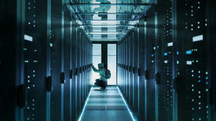 Fototapeta premium A Hooded Hacker With Laptop Connects to Rack Server and Steals Information from Corporate Data Center.