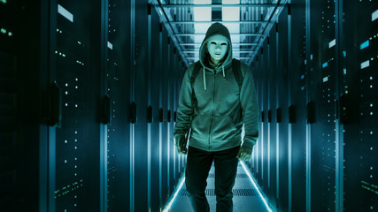 Shot of a Masked Hacker in a Hoodie Standing in Data Center with Rows of Rack Servers by His Sides.