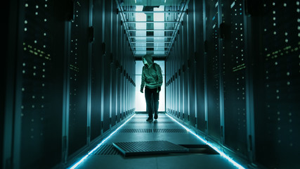 Hooded Hacker in a Mask Walks Through Working Data Center with Open Floor Hatch in the Middle of it.