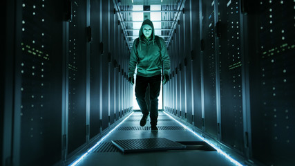 Hooded Hacker in a Mask Walks Through Working Data Center with Open Floor Hatch in the Middle of it.