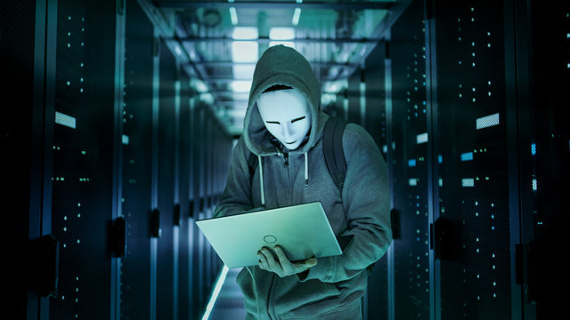 Mid Shot of a Masked Hacker in a Hoodie Standing in the Middle of Data Center full of Rack Servers and Hacking it with His Laptop.