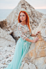 Fototapeta na wymiar Young beautiful red-haired woman in a luxurious dress stands on a rocky shore of the Adriatic Sea among large stones, close-up