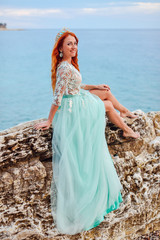 Young beautiful red-haired woman in mint dress sits on a large stone on the shore of the Adriatic Sea