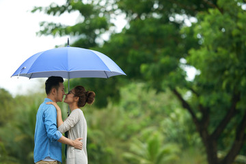 Side view portrait of young Asian couple kissing in rain, standing under umbrella, copy space