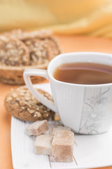 White with a flower pattern cup with tea, pieces of brown sugar and homemade cookies with cereals on a white saucer on a warm pastel orange background, a vertical frame