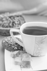 White cup with tea, slices of brown sugar and homemade cookies with cereals on a saucer against a background of a blurry basket with cookies, black and white frame