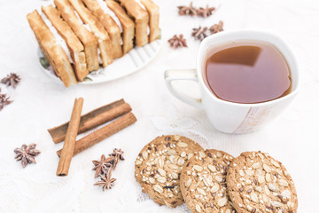 White cup with tea, homemade cookies with cereals, Viennese waffles with cream filling, cinnamon sticks, anise stars on a white lace tablecloth
