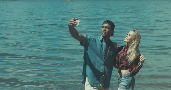 Young adult interracial couple taking a selfie on a beach, Caucasian female and African American man in casual clothes enjoying summer day